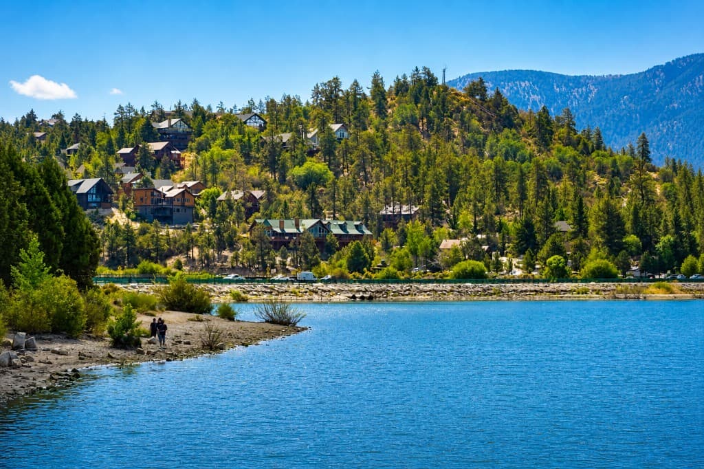 Calm mountain lake with a rocky shoreline, pine-covered hillside, and large homes among the trees under a clear sky.