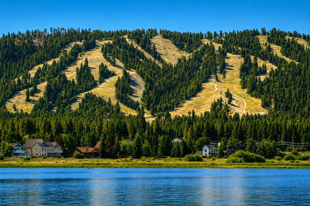 Mountain lake in the foreground, shoreline homes among pines, and a forested hillside with ski runs under a clear sky.